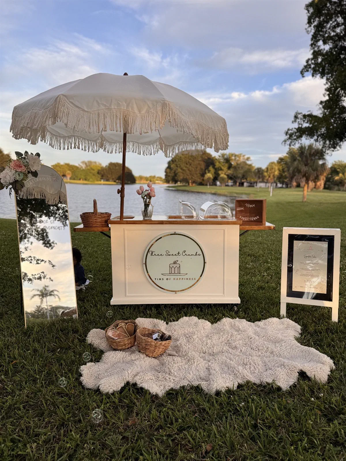 The Three Sweet Crumbs cake cart set up by a lake, with a fringed umbrella, pink tulips and a menu sign